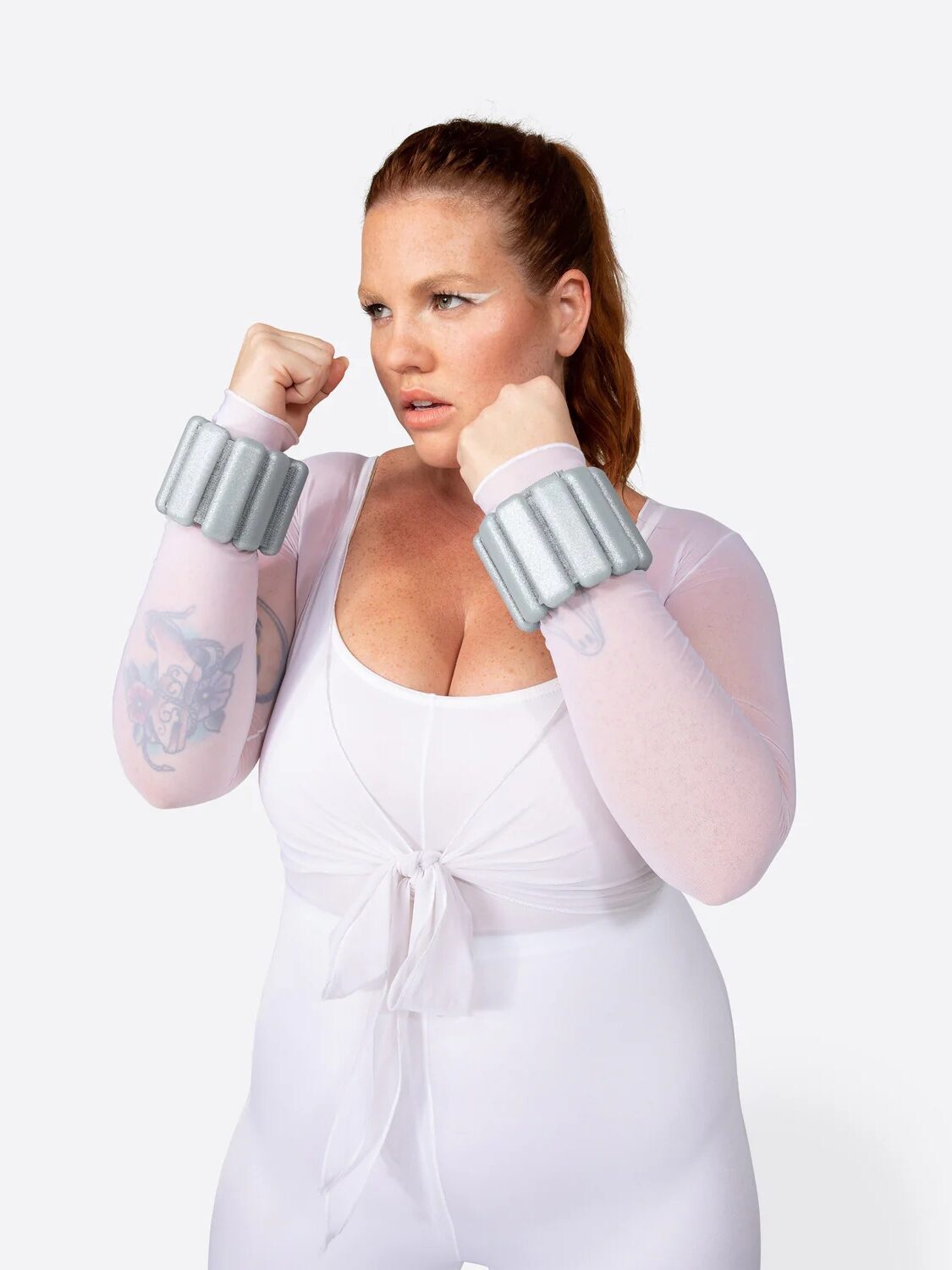 A person in a white outfit poses with fists raised, wearing silver wrist weights, ready to showcase the power of their training routine enhanced by massage guns.