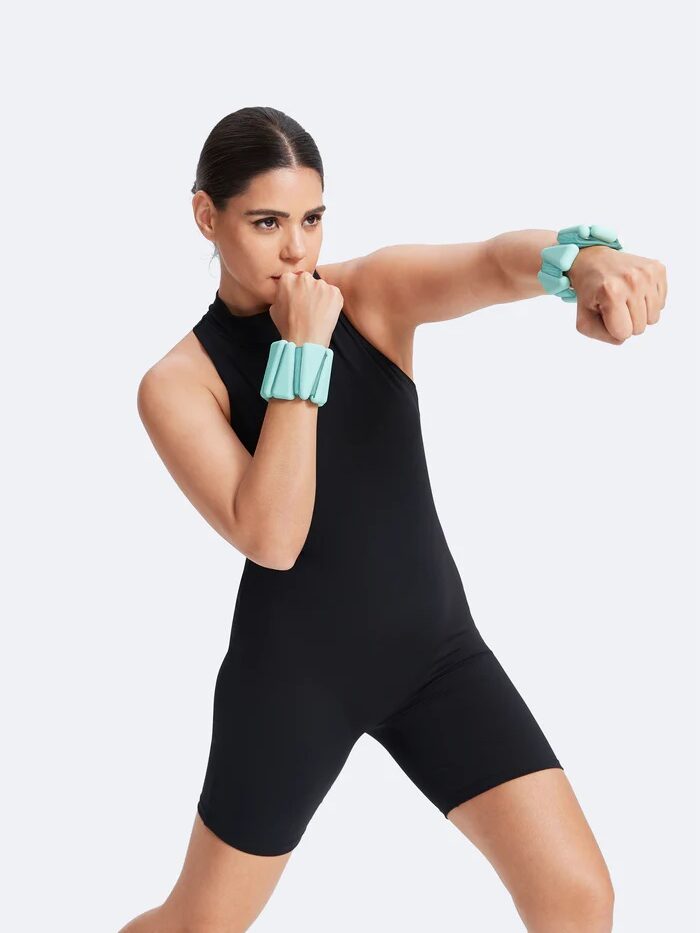 A person in a black athletic outfit strikes a boxing pose with teal wrist weights, embodying strength and focus. Nearby, massage guns lie ready to soothe tired muscles against the neutral backdrop.