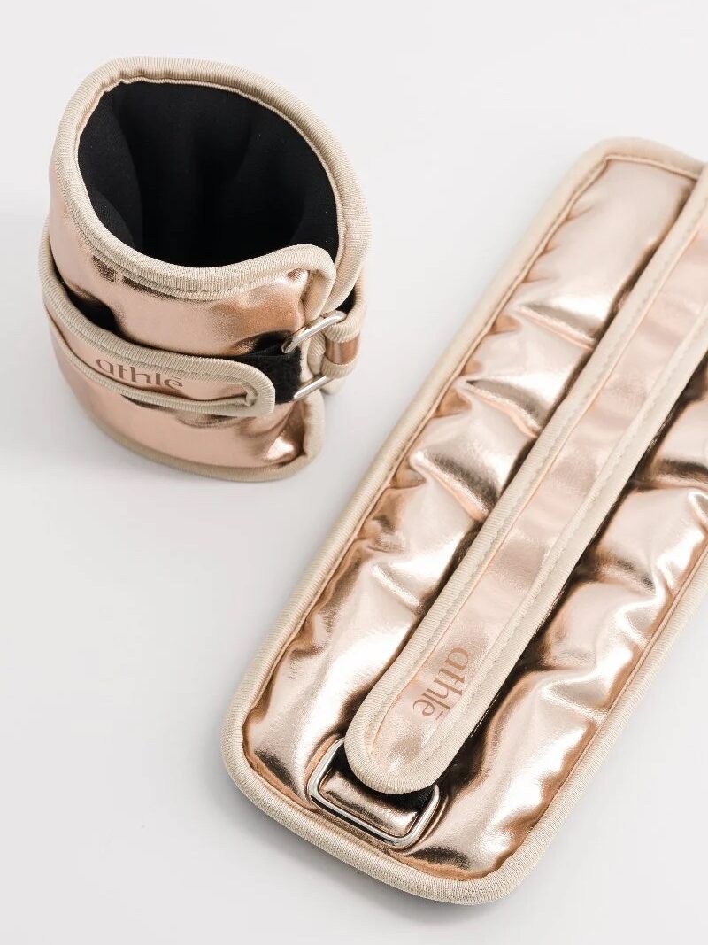 Rose gold ankle weights elegantly arranged on a white background, with one standing upright and the other lying flat, reminiscent of the sleek design often seen in premium massage guns.