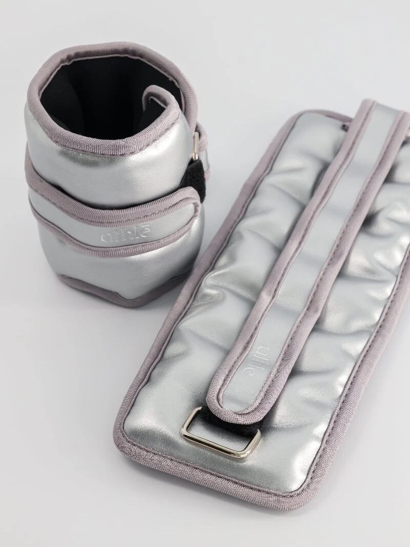 Silver, cushioned ankle weights on a white background; one weight stands upright while the other lies flat, showcasing its adjustable strap. Perfect for enhancing workouts, these pair seamlessly with your favorite fitness tools like massage guns.