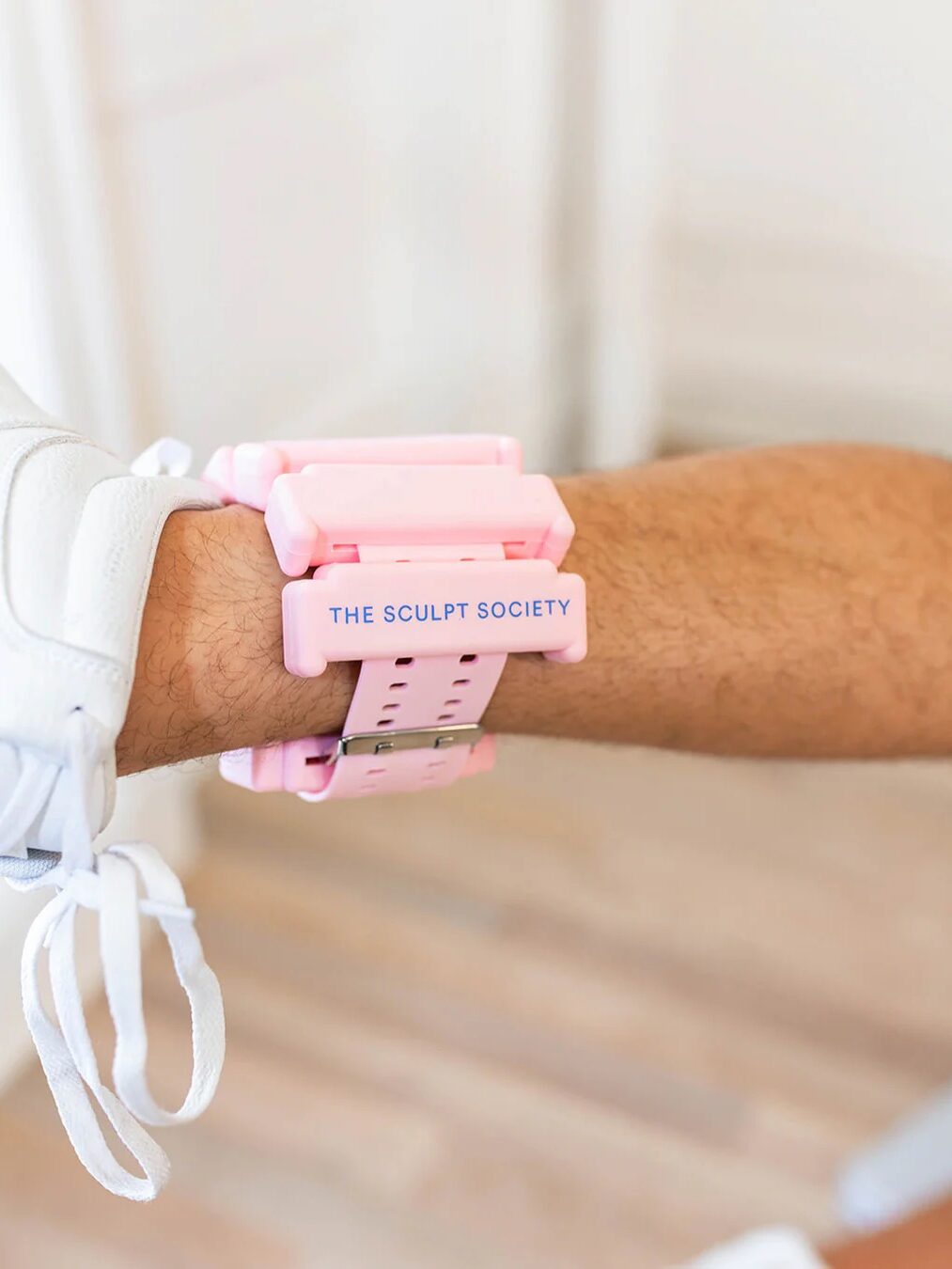 A pink fitness wristband reading "The Sculpt Society" graces a person's wrist, subtly complemented by part of a sneaker in view. Nearby, massage guns rest, promising to ease post-workout tension.