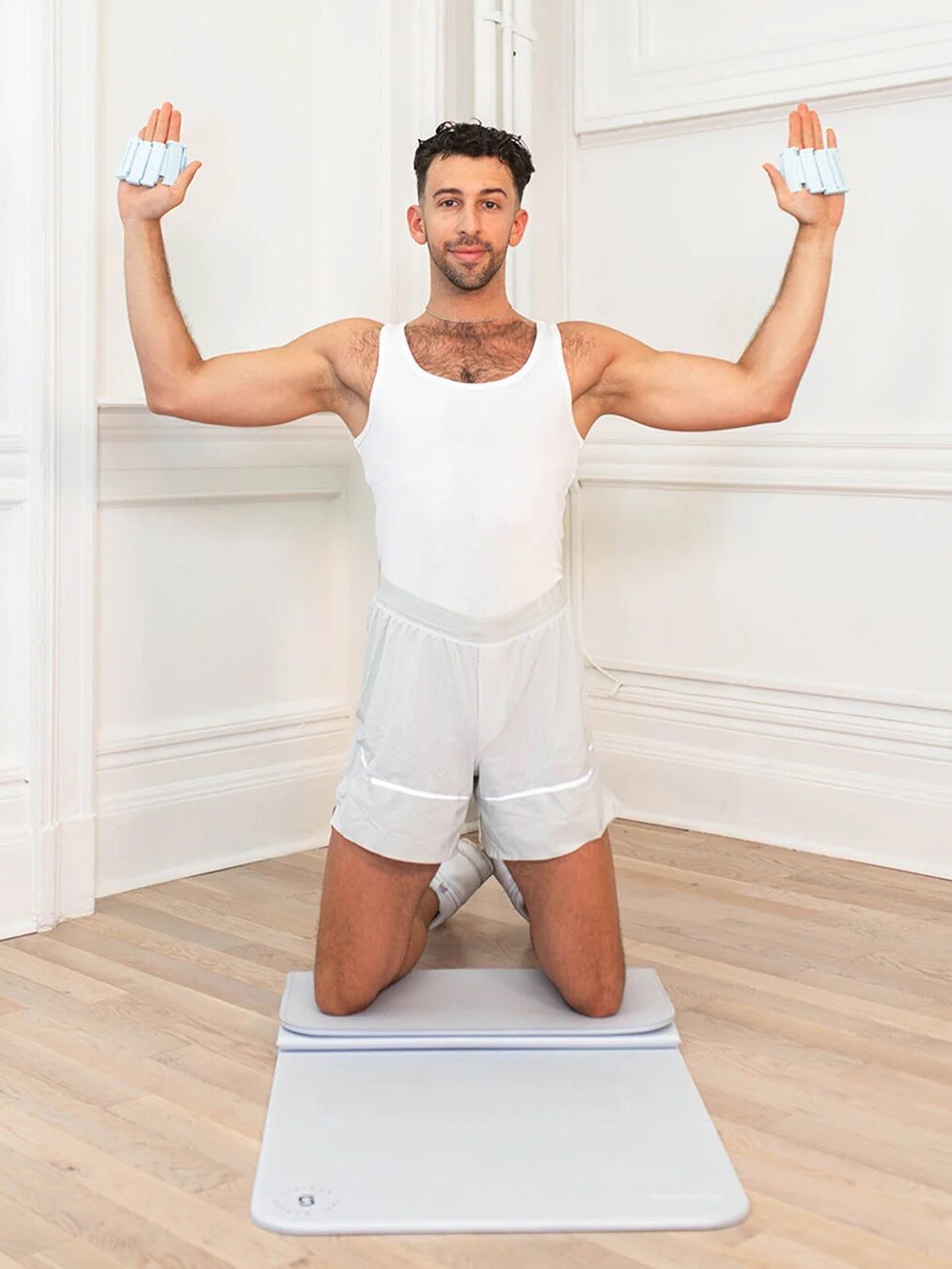 Person kneeling on an exercise mat, wearing a white tank top and shorts, holding weights in both hands raised in a W shape, as a massage gun sits nearby ready to soothe post-workout muscles.