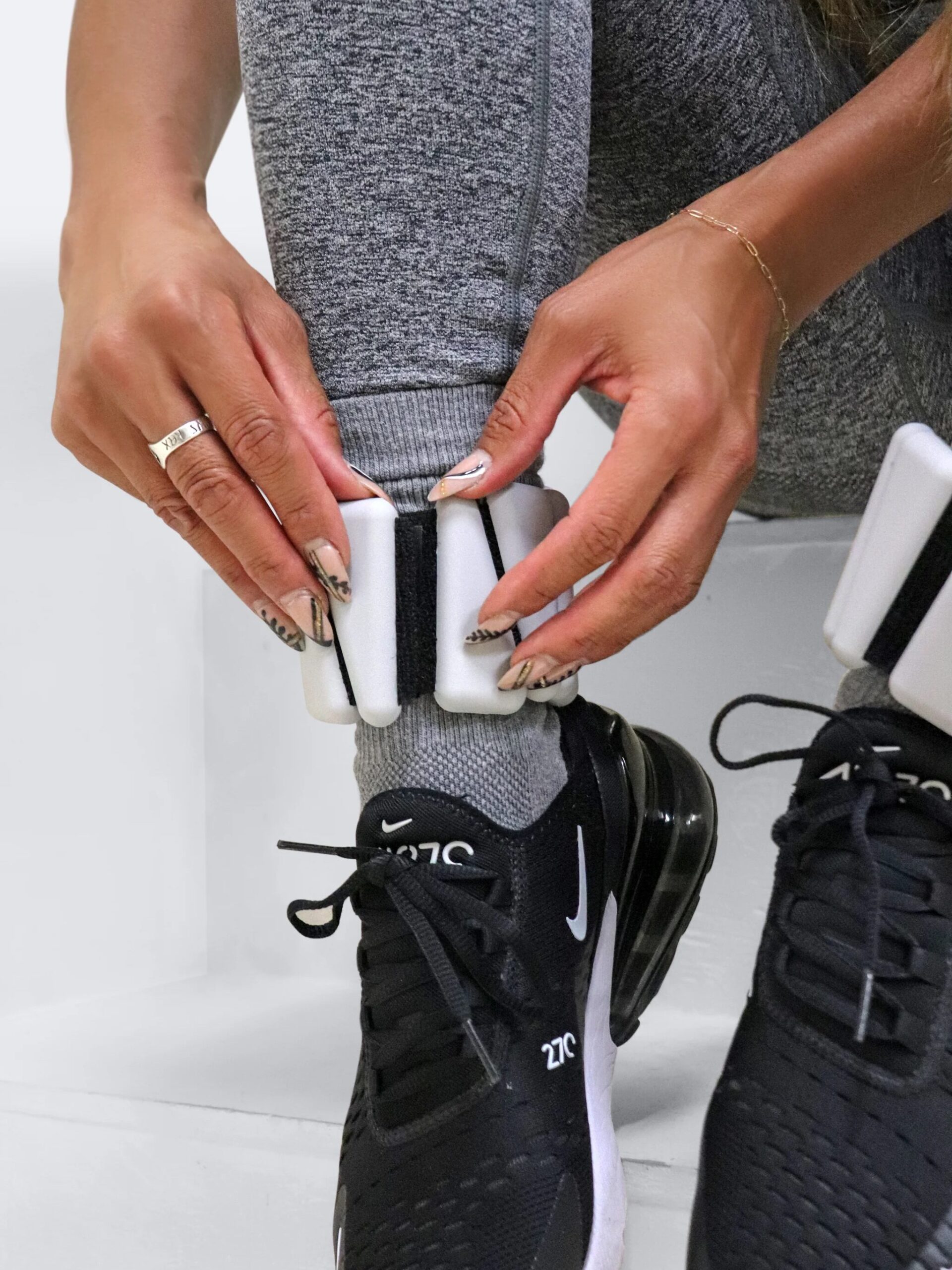 A person wearing gray leggings adjusts a white ankle weight over their sock, while a black athletic shoe rests nearby. In the background, a massage gun sits ready for post-workout recovery.