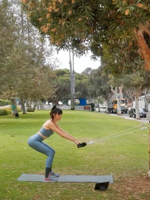 A person is exercising with resistance bands attached to a tree in a tranquil park, enjoying the fresh air and planning to unwind later with their massage gun for optimal muscle recovery.