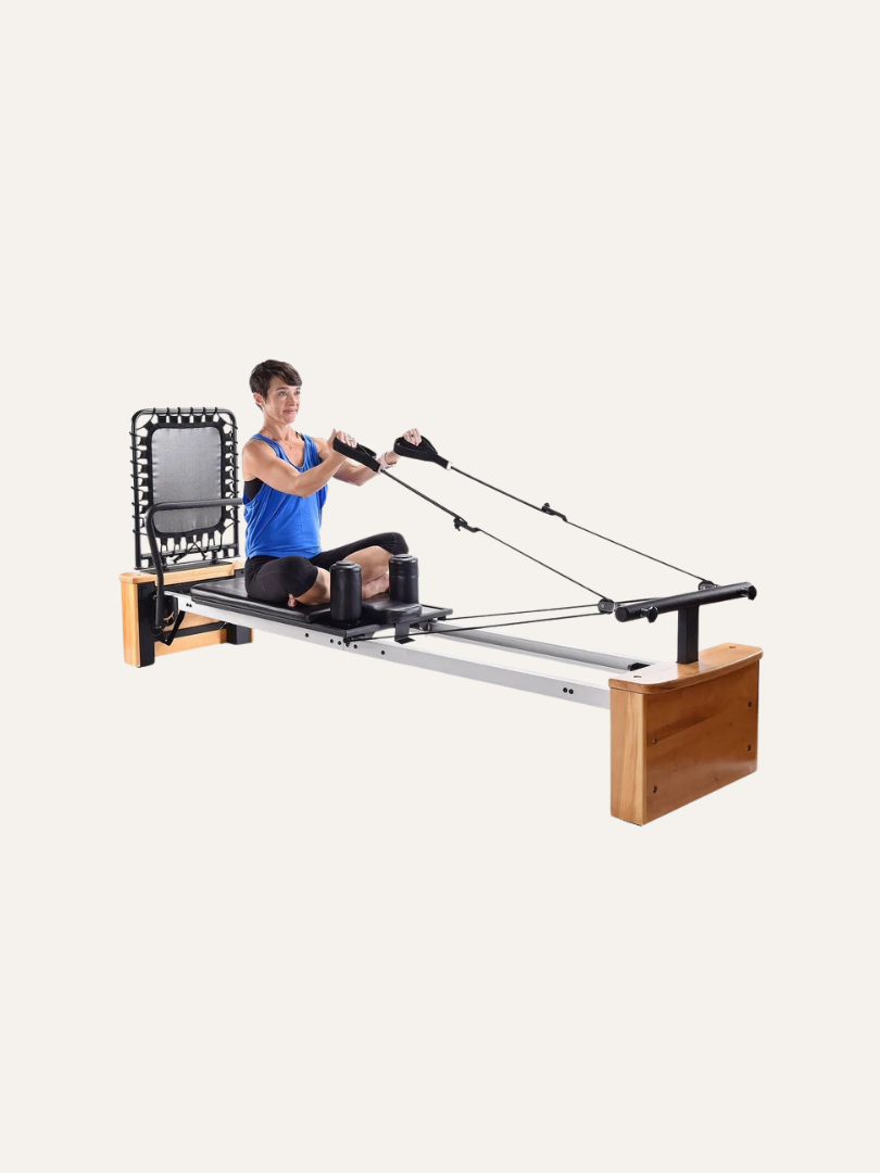 A person engages with a Pilates reformer machine, pulling cables while seated with legs extended and knees slightly bent. After the session, they reach for their massage guns to soothe their muscles and enhance post-workout recovery.