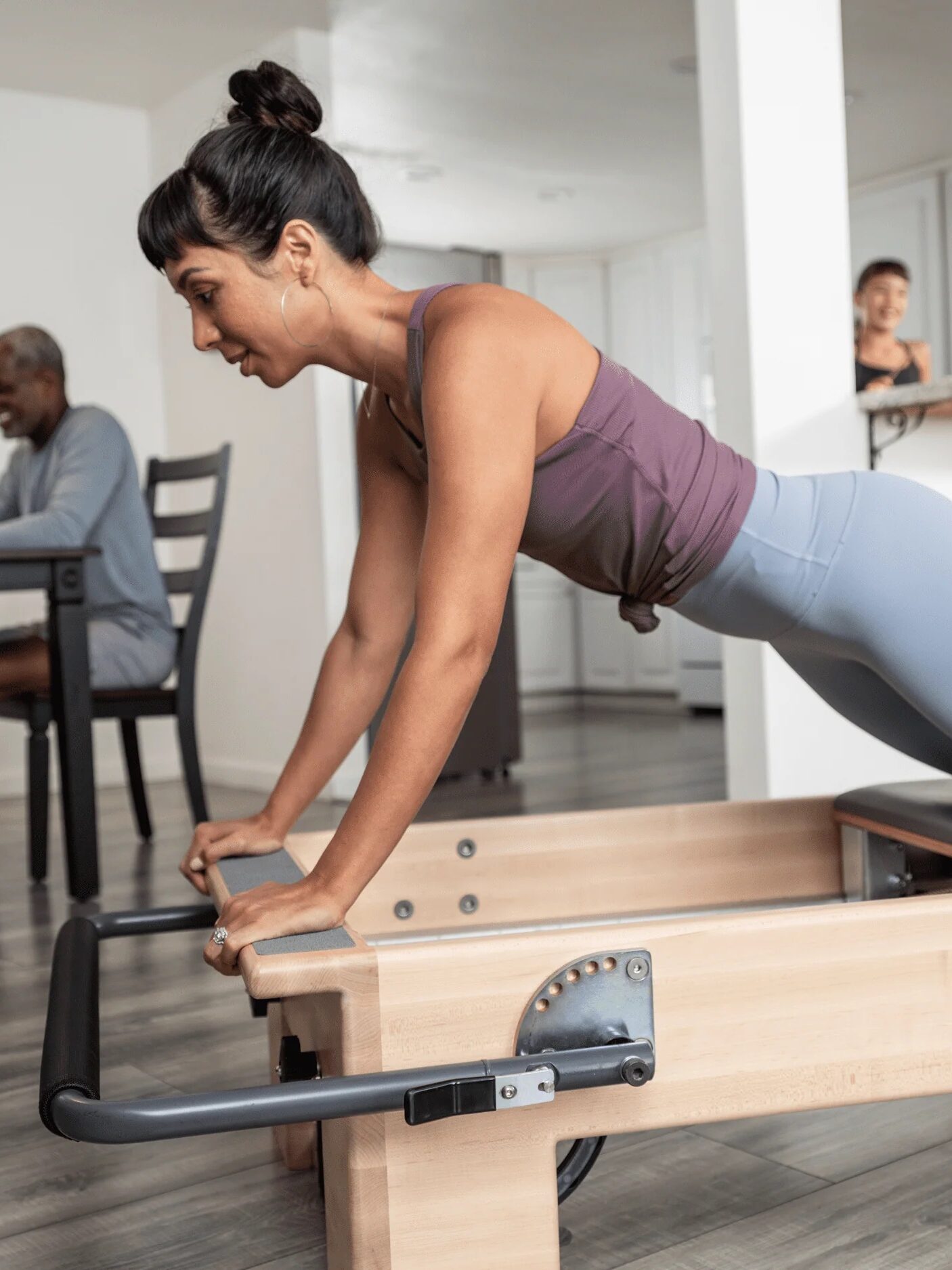 A woman exercises on a Pilates reformer in a living room while two people chat at the kitchen counter, occasionally glancing at a massage gun brochure. Meanwhile, a man sits at the dining table using his laptop.