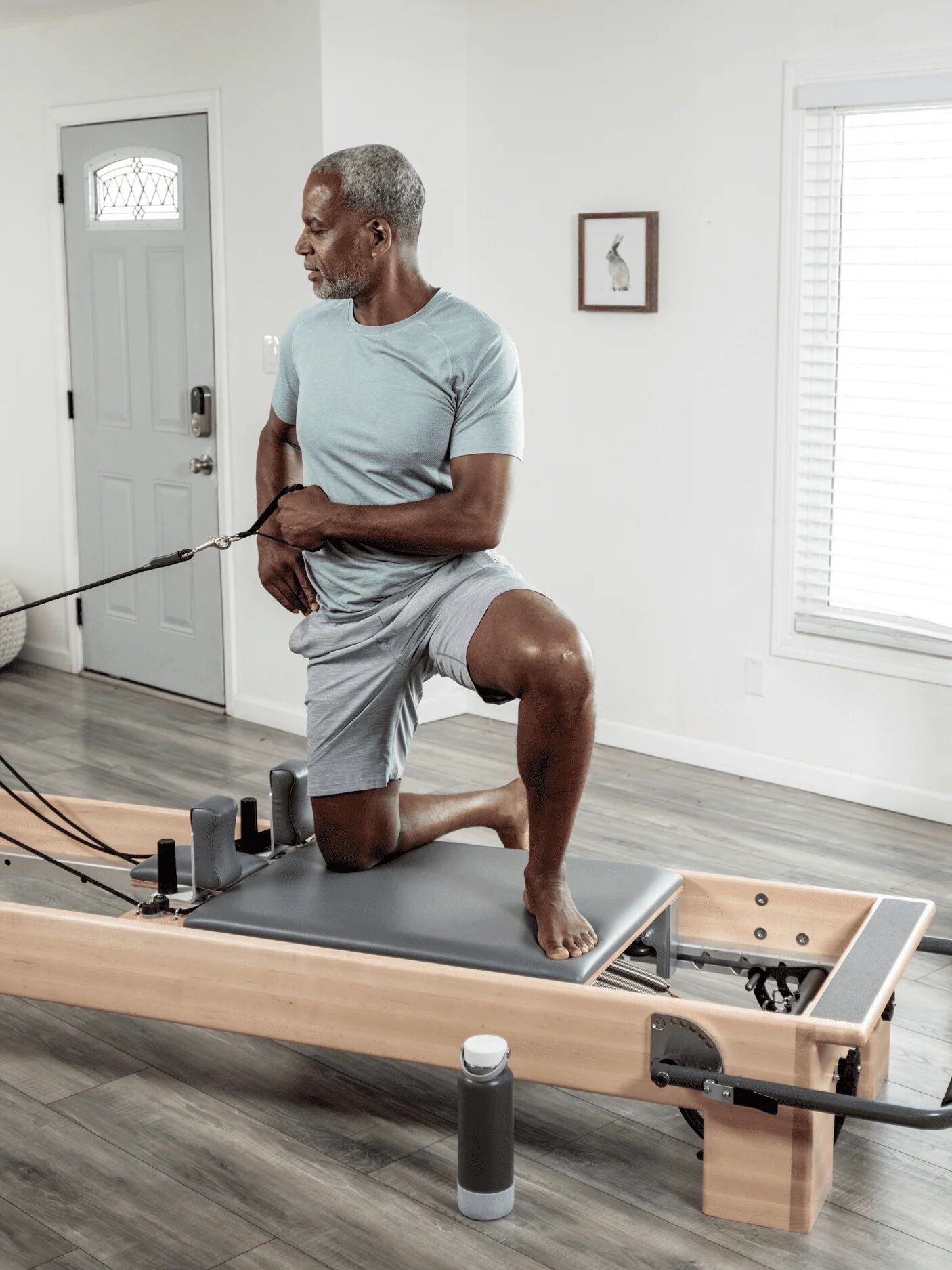 An older man in a light blue outfit performs exercises on a Pilates reformer machine next to a brightly lit room with a sofa, painting, and potted plant, considering the addition of massage guns to his routine for enhanced muscle recovery.