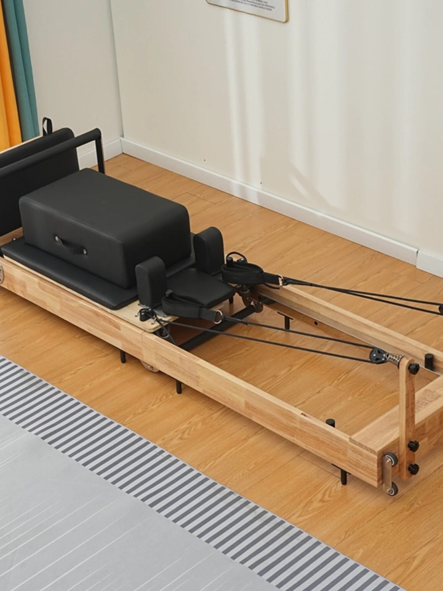 A wooden Pilates reformer with black cushioning and adjustable ropes sits on a wooden floor near a white wall. A small potted plant adds charm, while a set of sleek massage guns rests nearby, promising ultimate relaxation after each session.