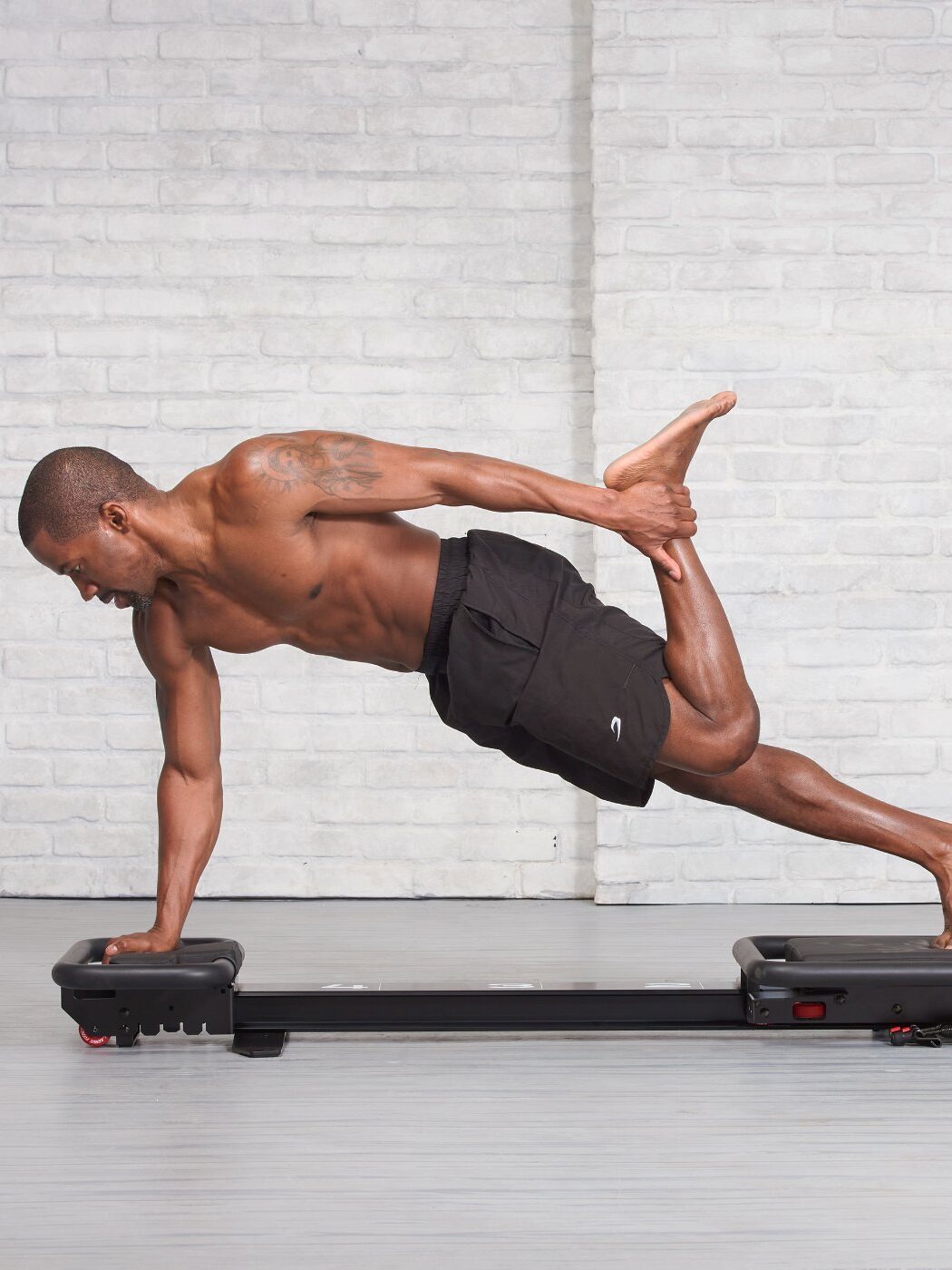 A person skillfully performs a side plank with one hand, while balancing on fitness equipment against a gray brick wall. Nearby, massage guns rest, hinting at the recovery routine that follows this impressive hold.