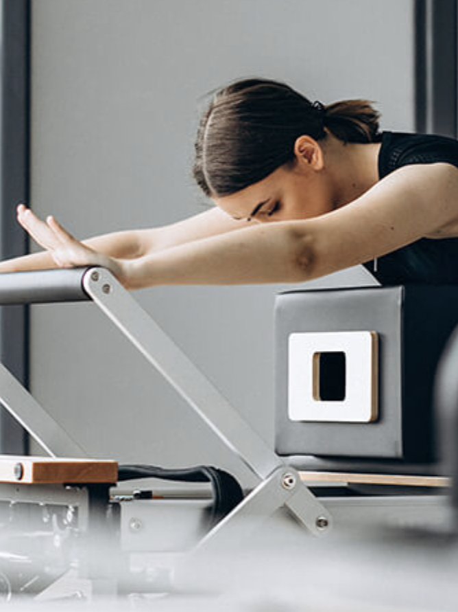 A person performs Pilates on a reformer machine, leaning forward with arms extended, feeling the stretch deepen, while a massage gun rests nearby for post-workout relaxation.