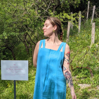 A person in a blue dress stands next to a sign in a garden, displaying a tattoo on one arm.