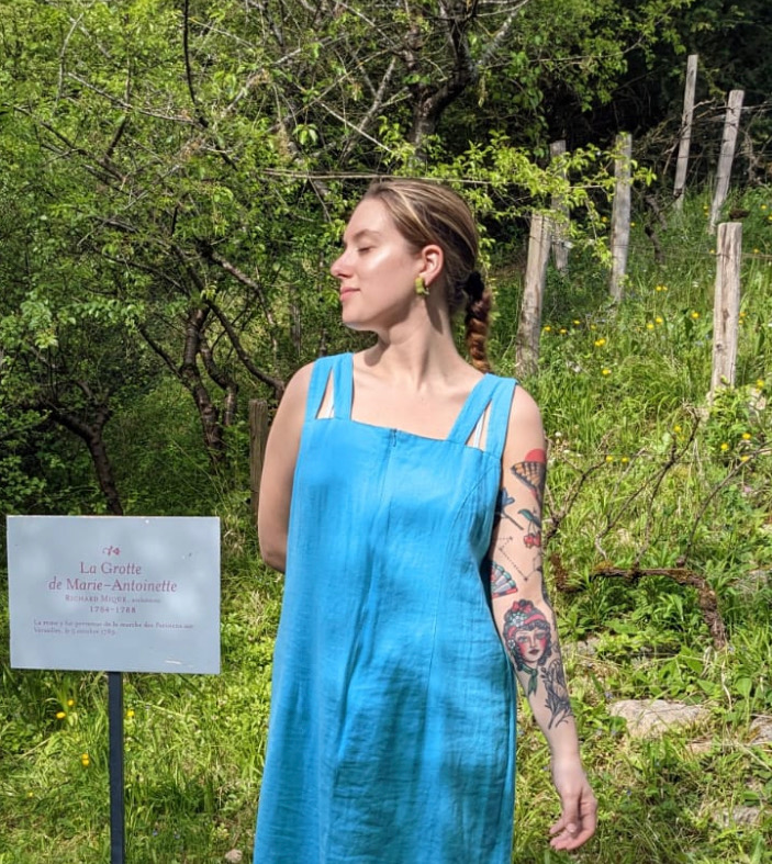 A person in a blue dress stands next to a sign in a garden, displaying a tattoo on one arm.