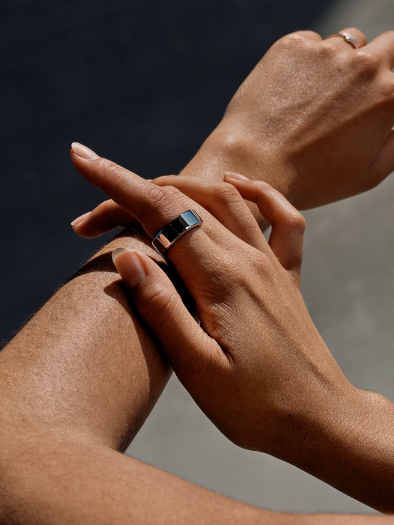 Two hands with rings, including a sleek Oura Ring, gracefully overlap against a neutral background. The focus is on the jewelry.