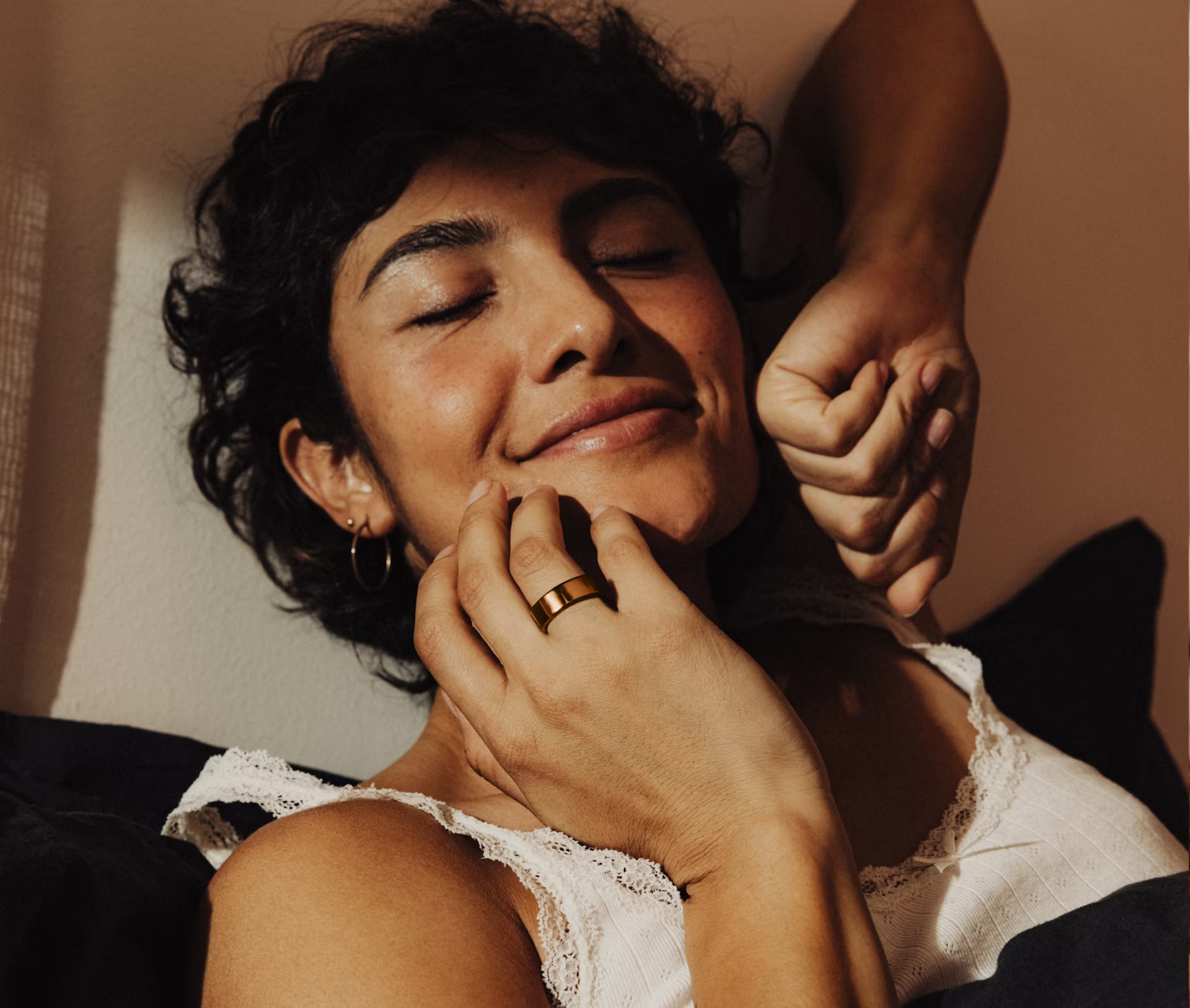 Person with short hair and wearing a white lace top smiles with eyes closed, resting on a pillow. An Oura Ring gleams softly on their finger, capturing the serenity of the moment.