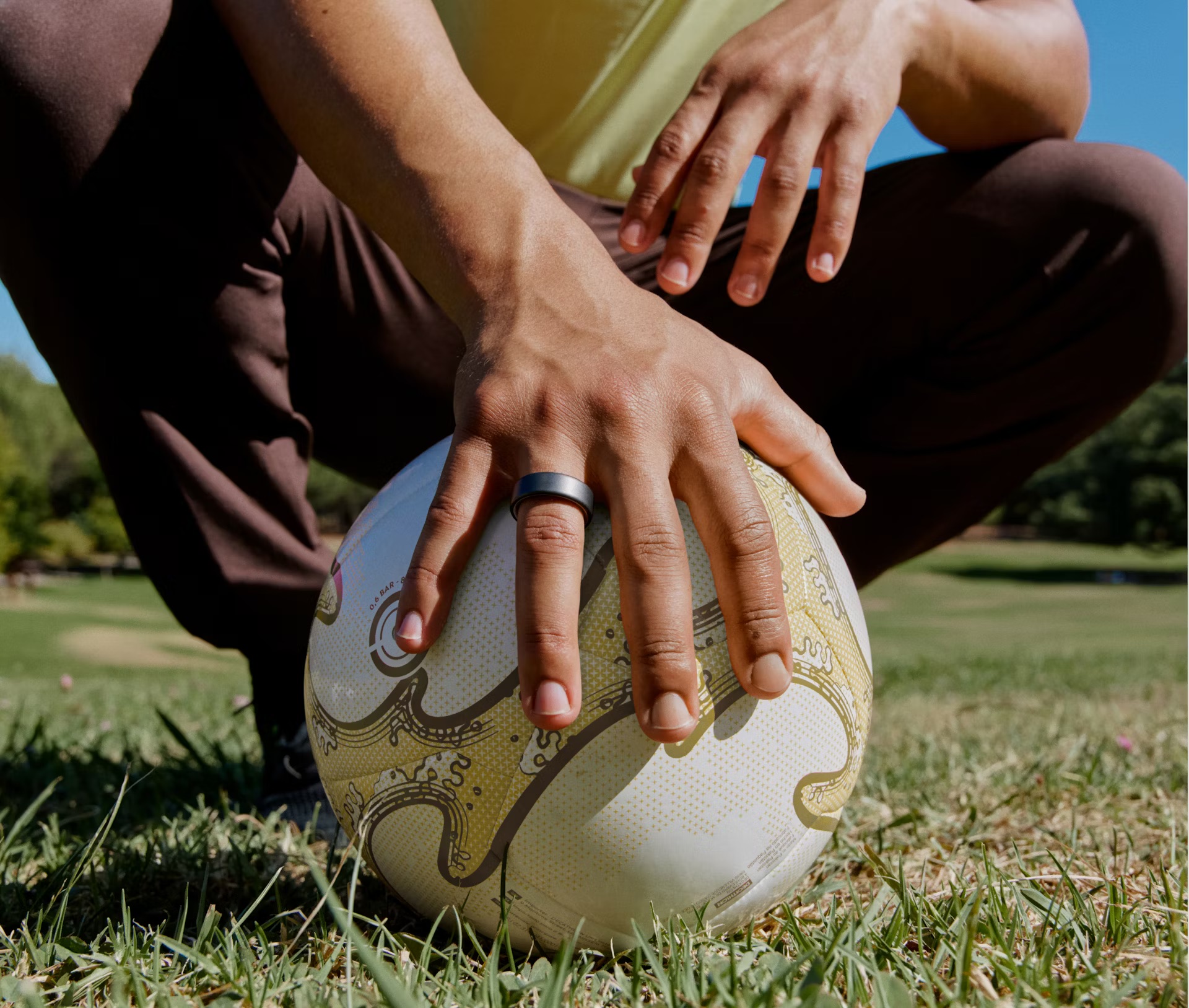 A person kneels on grass, sporting an Oura Ring while holding a soccer ball with one hand. Their other hand rests confidently on their knee.