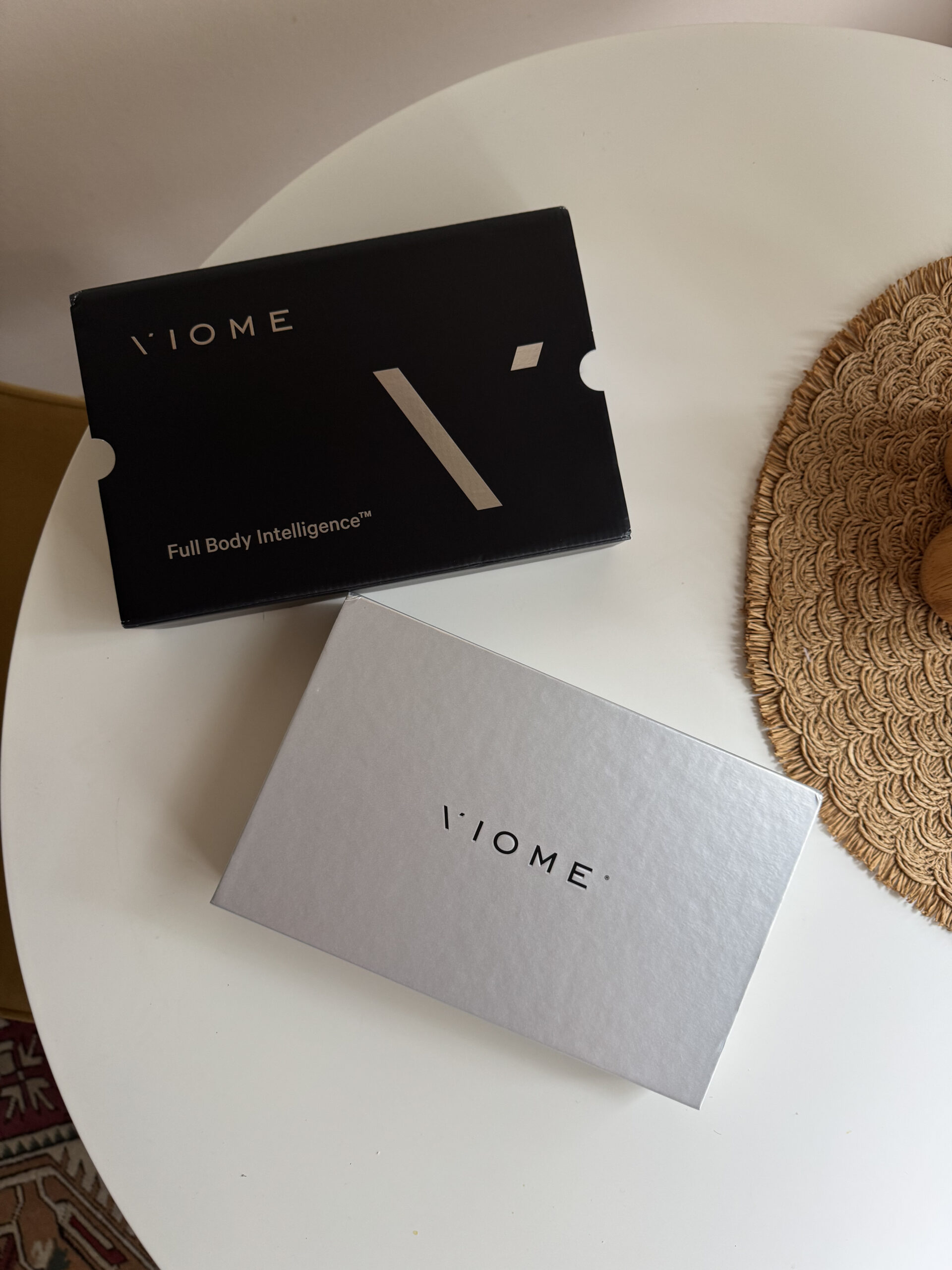 Two Viome product boxes, one black labeled "Full Body Intelligence" and one silver, are placed on a round white table. A woven placemat is partially visible beside them.