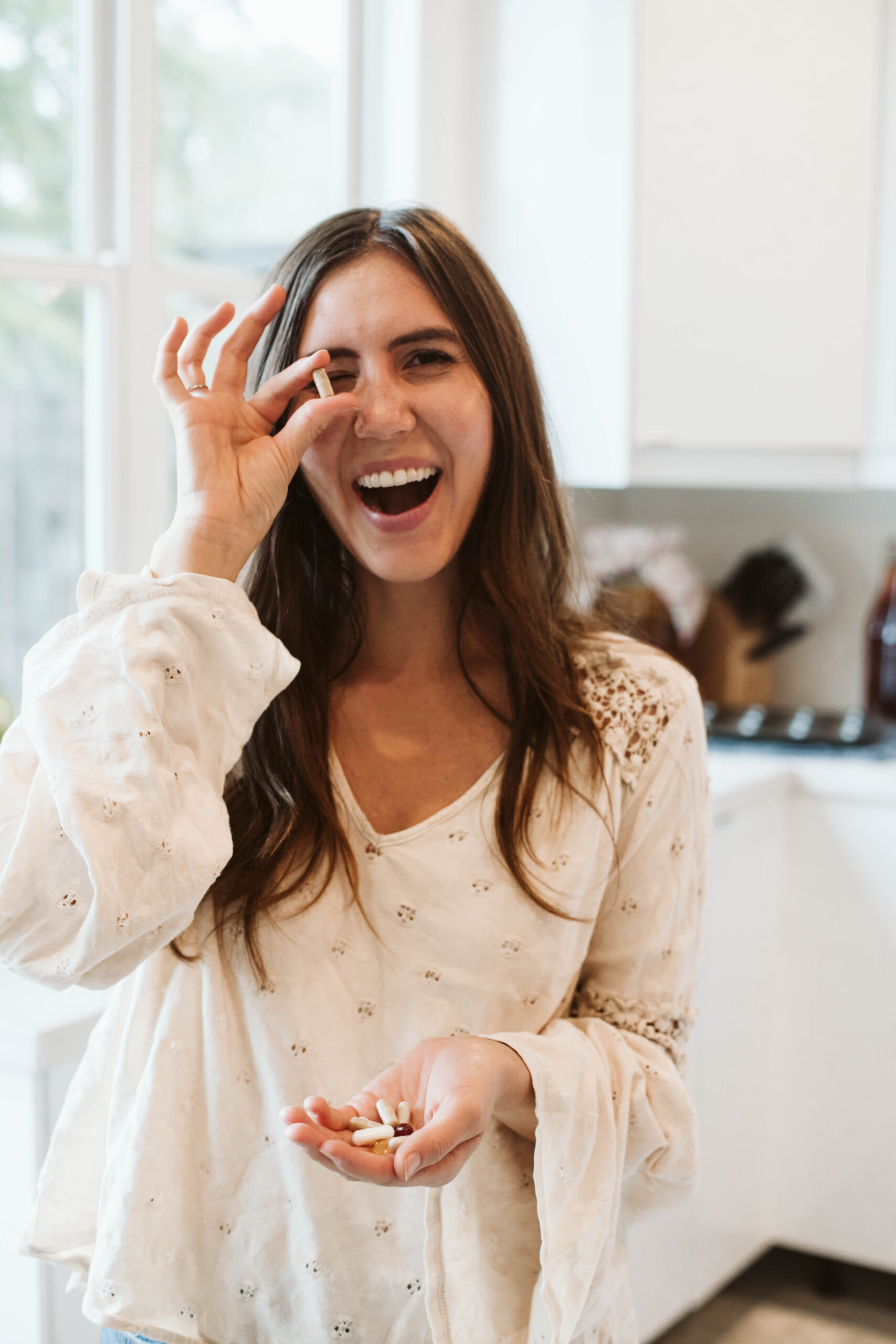 Woman in a kitchen smiling, holding several capsules in one hand, and playfully holding one up to her eye with the other hand.