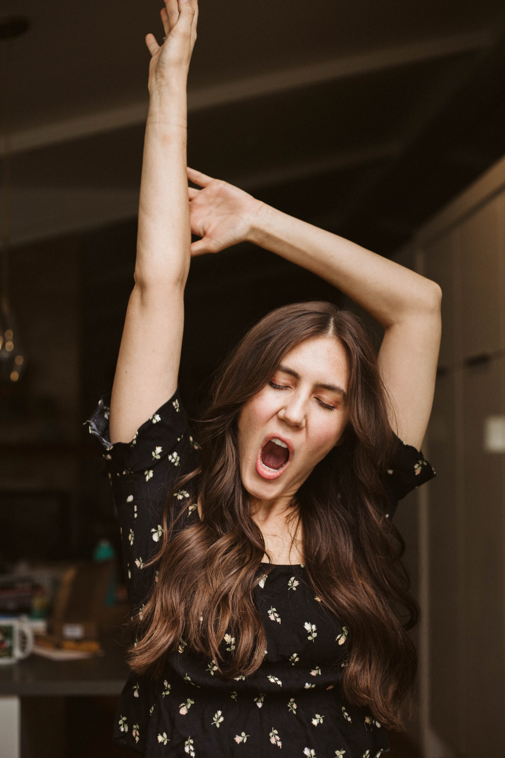 Woman with long hair yawning, stretching one arm overhead, wearing a floral dress indoors.