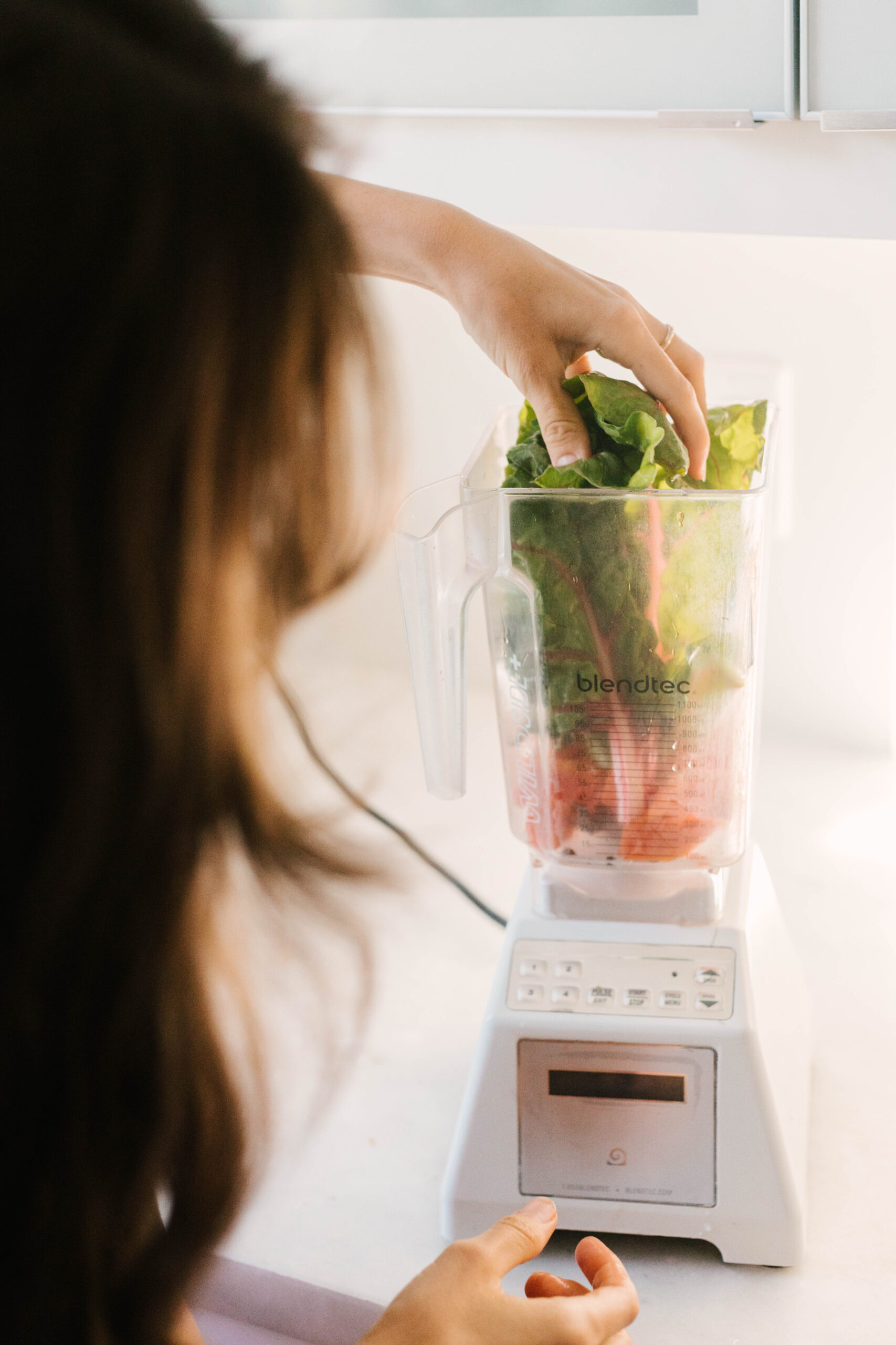 Person placing greens into a white blender filled with assorted vegetables.