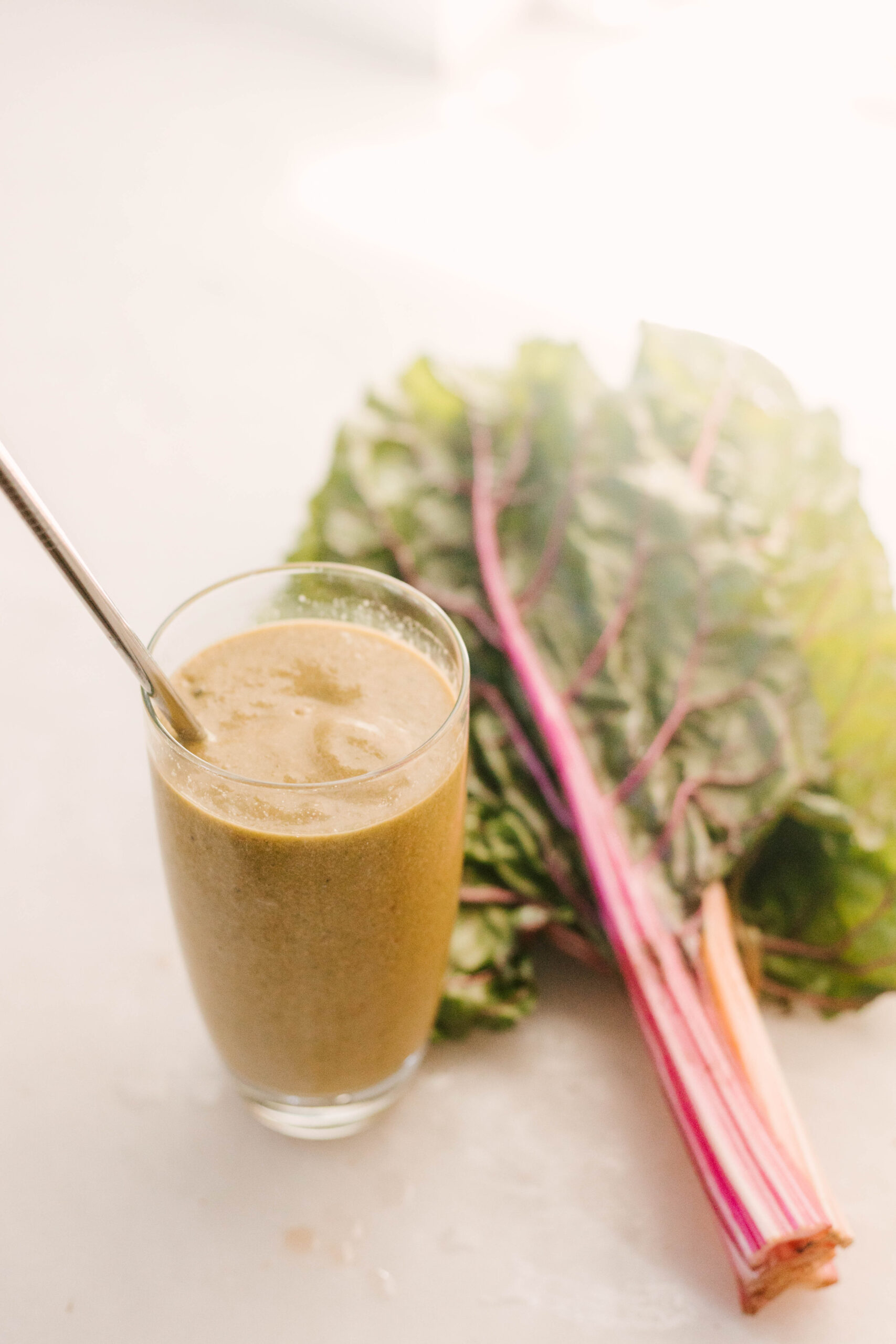 A green smoothie in a glass with a metal straw, placed next to a bunch of rainbow chard on a light surface.