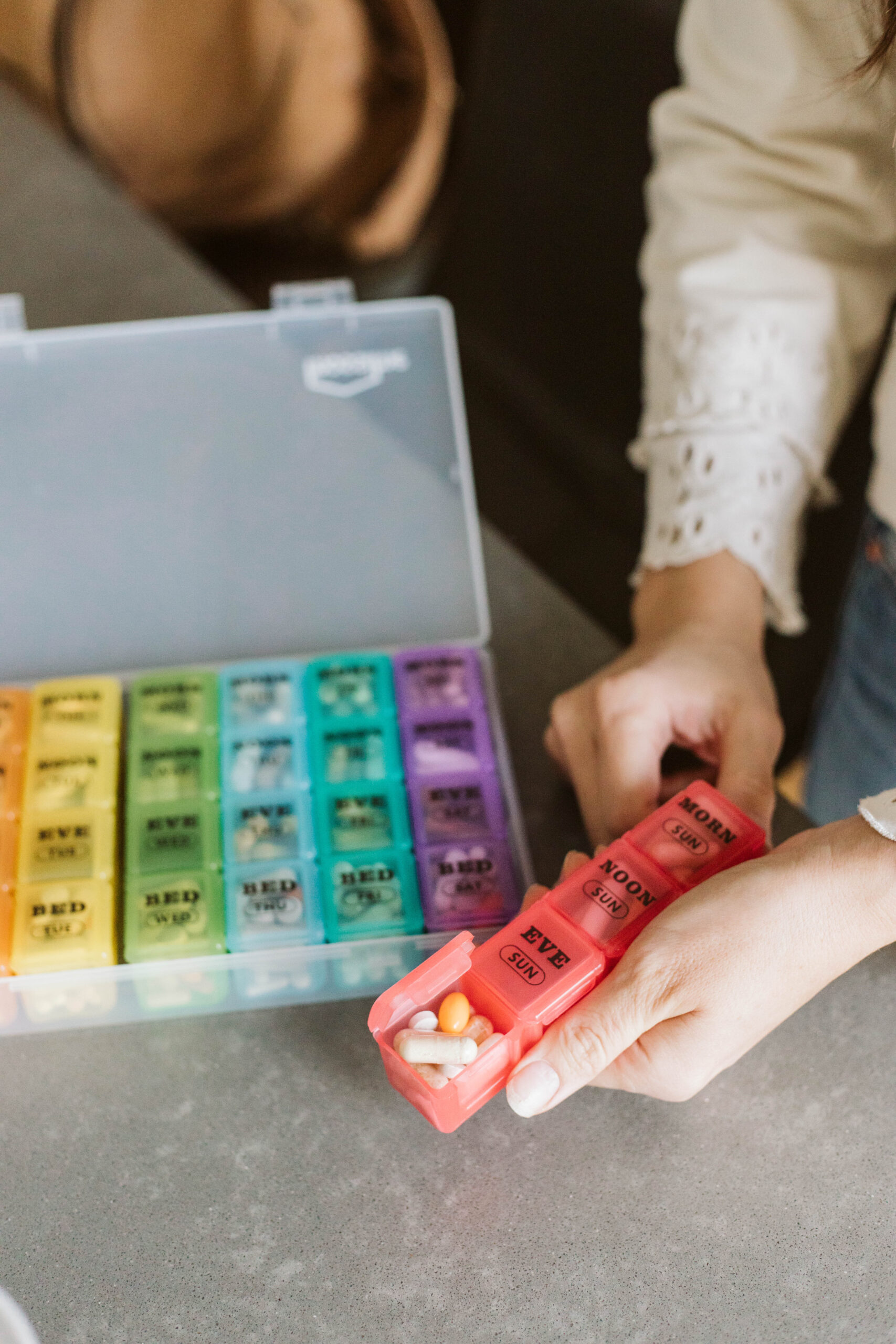 Person holding a red pill organizer labeled "NIGHT" with pills inside, part of a larger weekly pill organizer on a table.