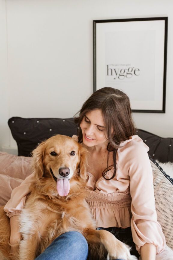 Woman sitting on a couch with a golden retriever, smiling. A framed print with the word "hygge" is on the wall behind them.