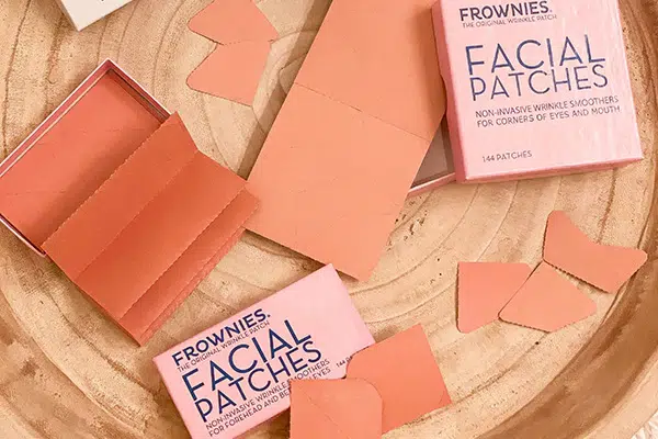 Two pink boxes of Frownies Facial Patches are open on a wooden surface, with several patches scattered around.