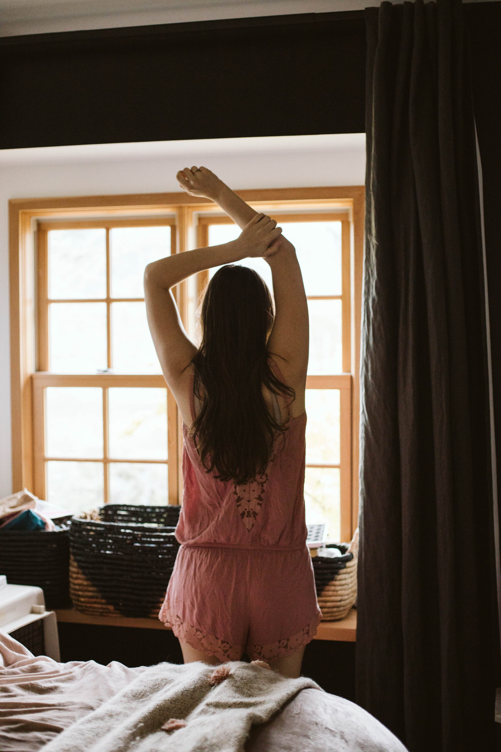 Person in pink pajamas stretches with arms raised, standing in front of a window with wooden frames.