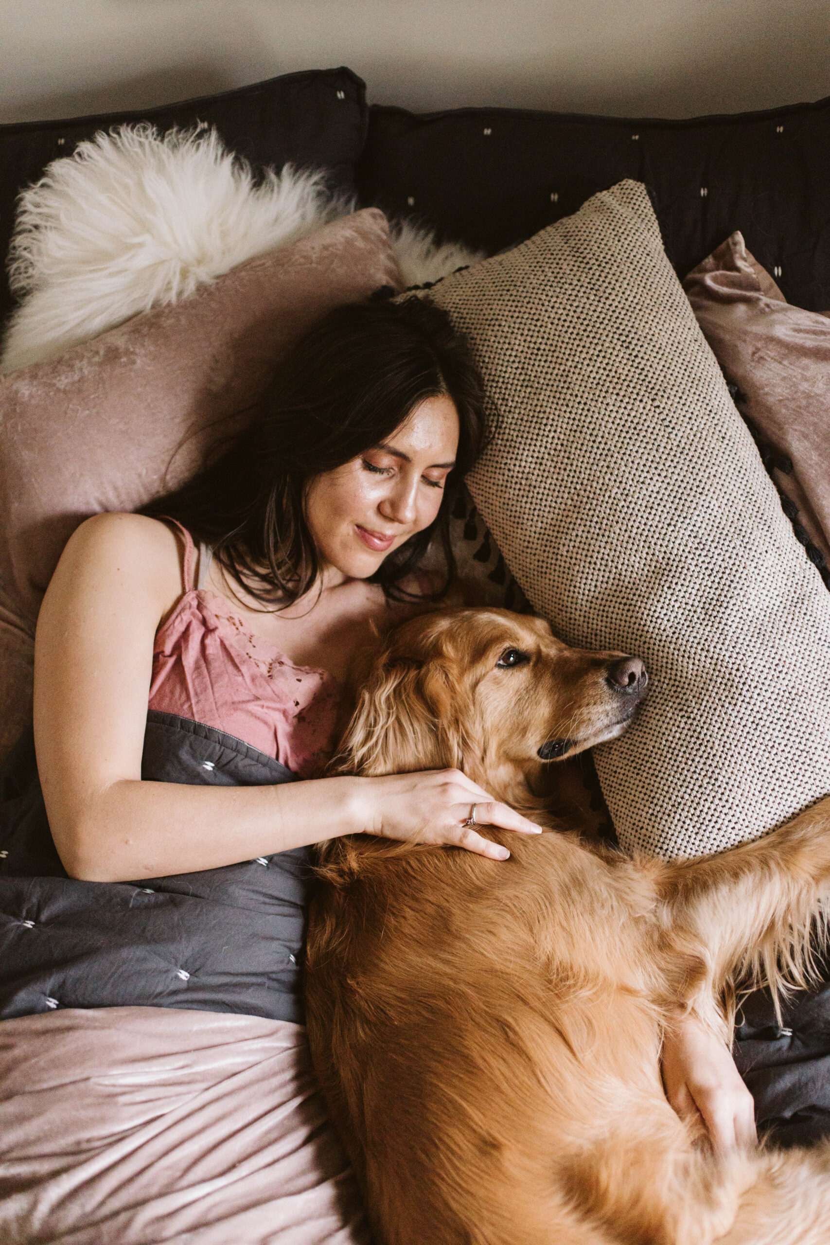 Woman lying on a bed, gently cuddling a Golden Retriever. The bed has soft pillows and a fluffy blanket.