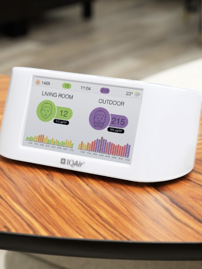 A digital air quality monitor sits on a wooden table, displaying indoor and outdoor air quality readings. The brand "IQAir" is visible on the device and in the corner of the image.