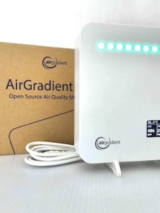 The White AirGradient air quality monitor, elegantly perched on a stand with its screen showcasing vital data, stands proudly in front of its box.