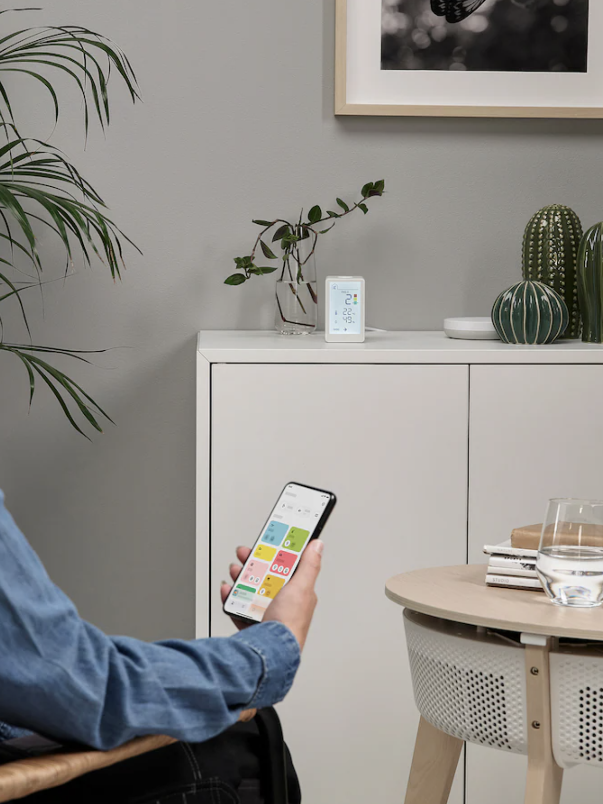 A woman in a denim jacket sits in a wicker chair, holding a smartphone and looking at the white cabinet.