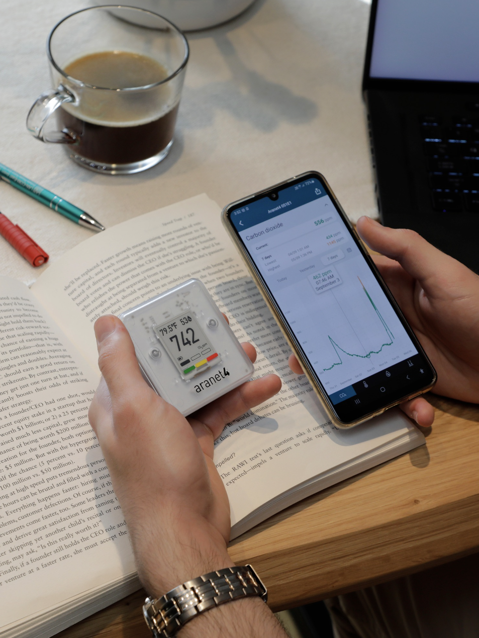 Person holds a phone displaying a graph and a digital monitor showing "742," while nearby, an open book, pens, and a cup of coffee are neatly arranged on the table.