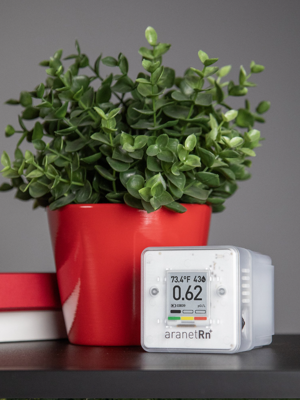 A red pot with a green plant sits beside a digital radon meter displaying 0.62.