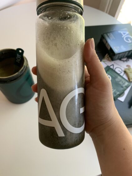 A hand holding a frothy drink in a clear bottle labeled "AG" with a matching jar and box in the background on a white surface.