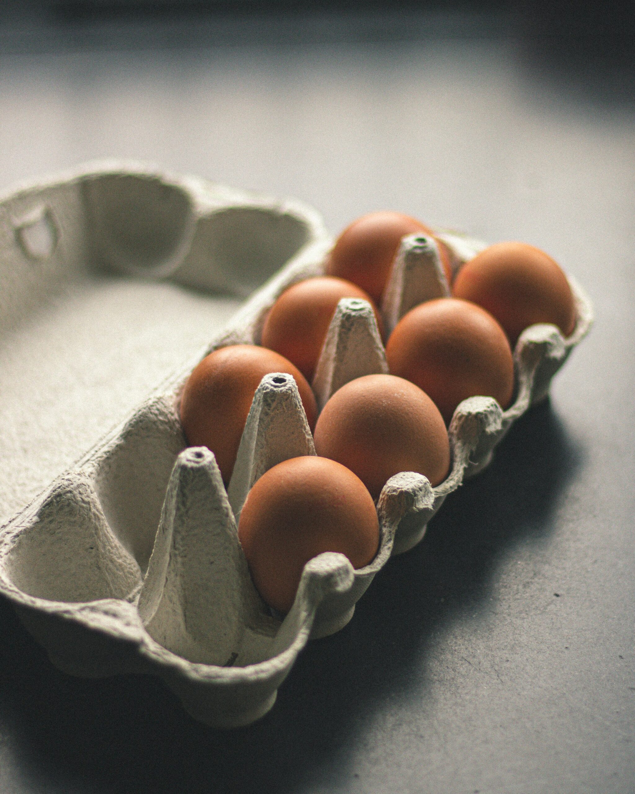 A carton with eight brown eggs sits on a dark surface, with four empty spaces in the container.