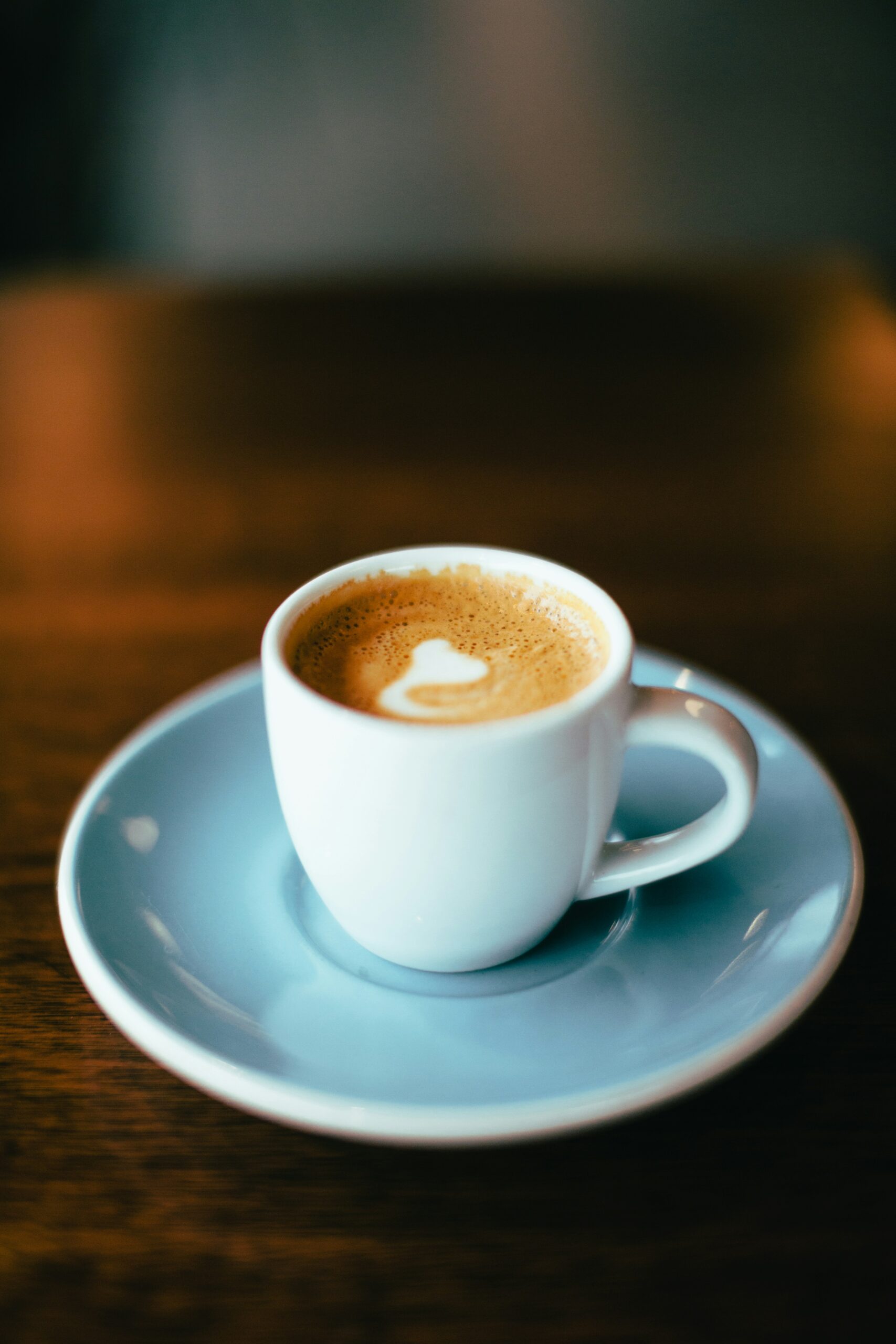 A white cup of coffee with a small amount of foam on top sits on a matching saucer on a wooden table.