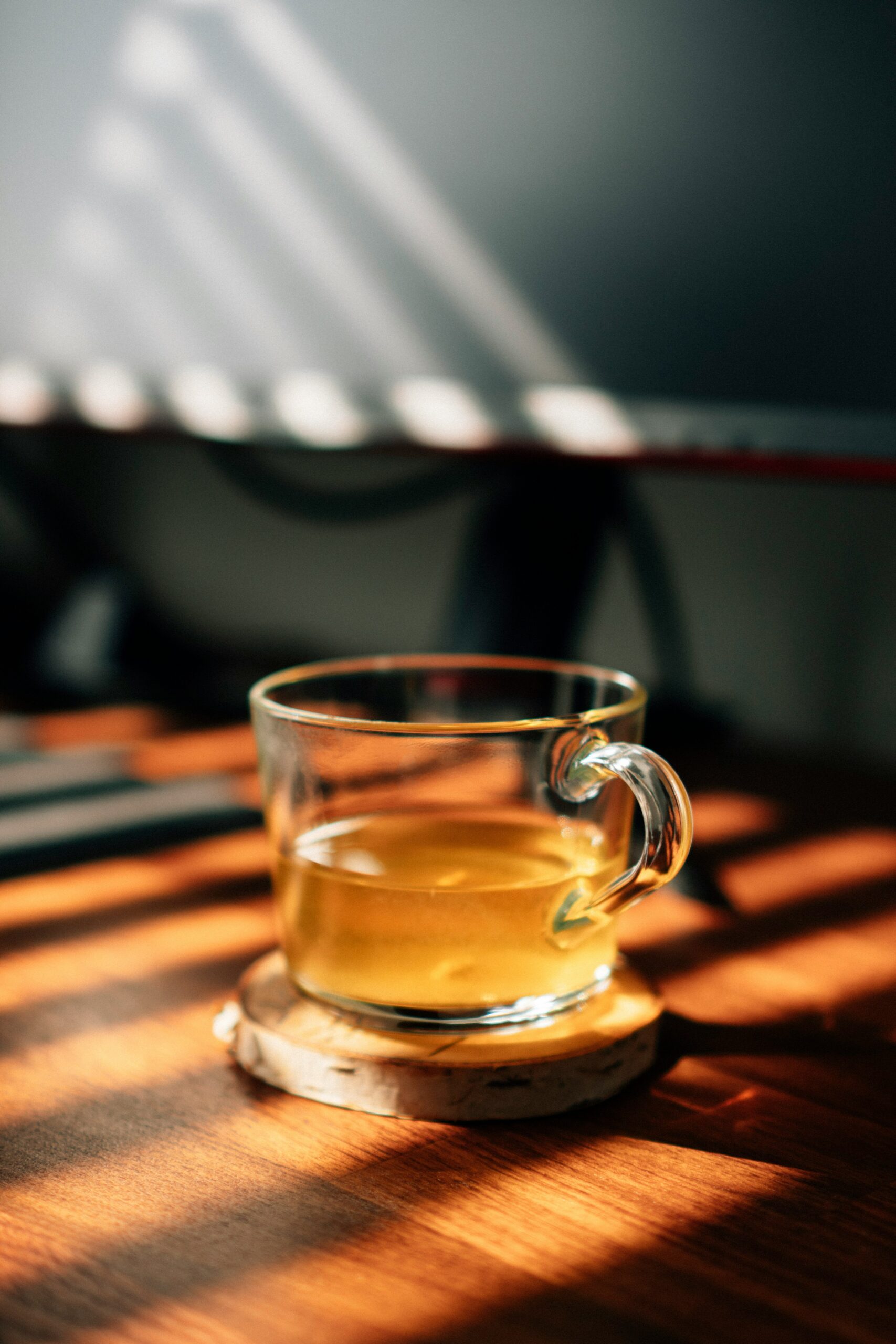 A glass cup of tea sits on a wooden table, illuminated by sunlight through blinds.