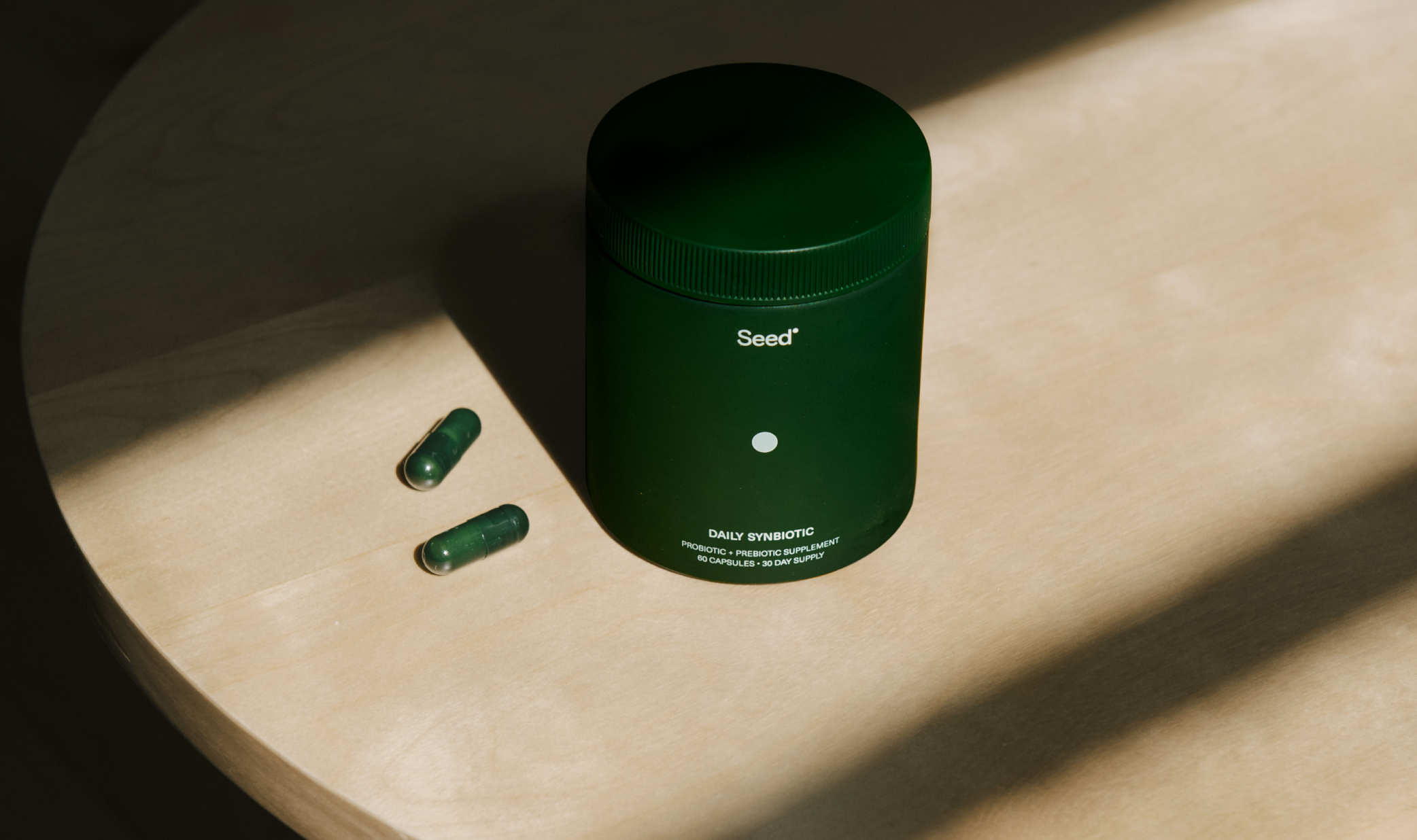 A green jar labeled "Seed" and two green capsules are placed on a wooden table with diagonal light shadows.