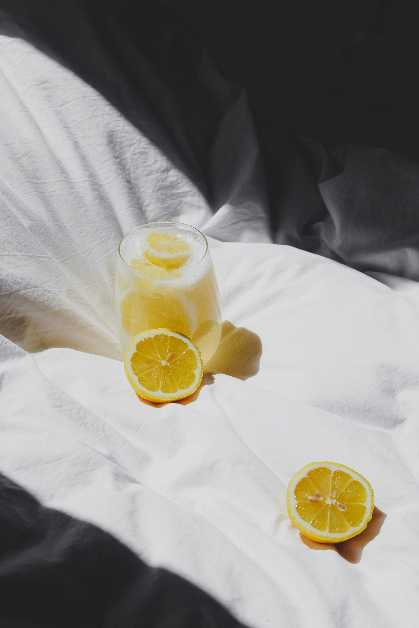 A glass of lemonade on a white fabric with two lemon halves nearby, illuminated by natural light.