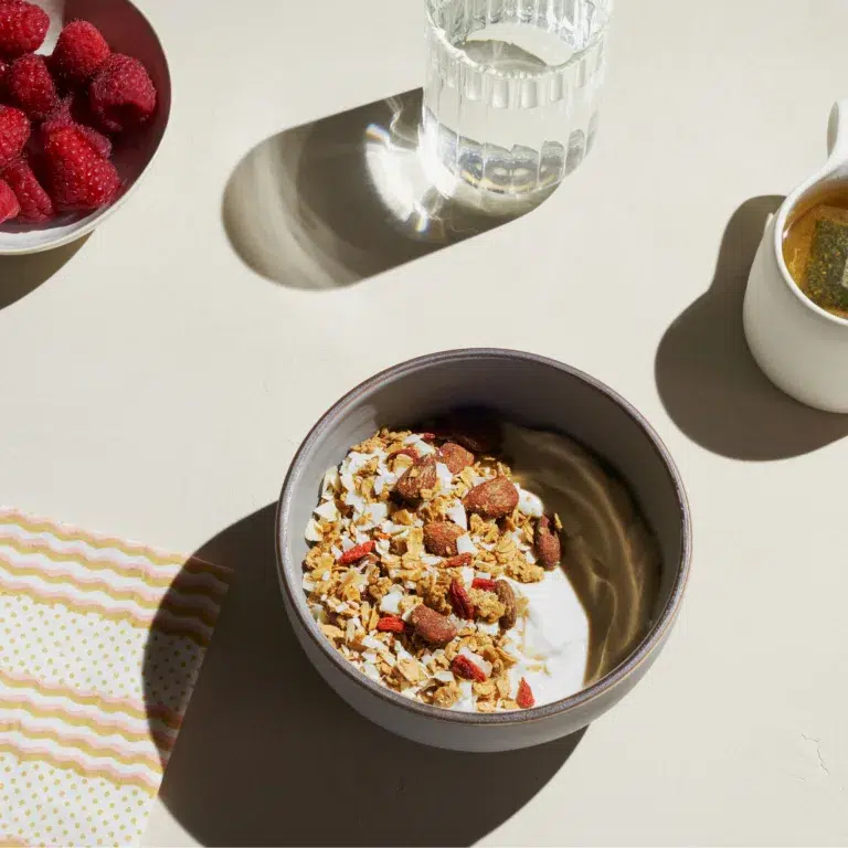 A bowl of yogurt topped with granola and almonds, accompanied by a glass of water, a cup of tea, and a bowl of raspberries on a beige table with a patterned napkin.