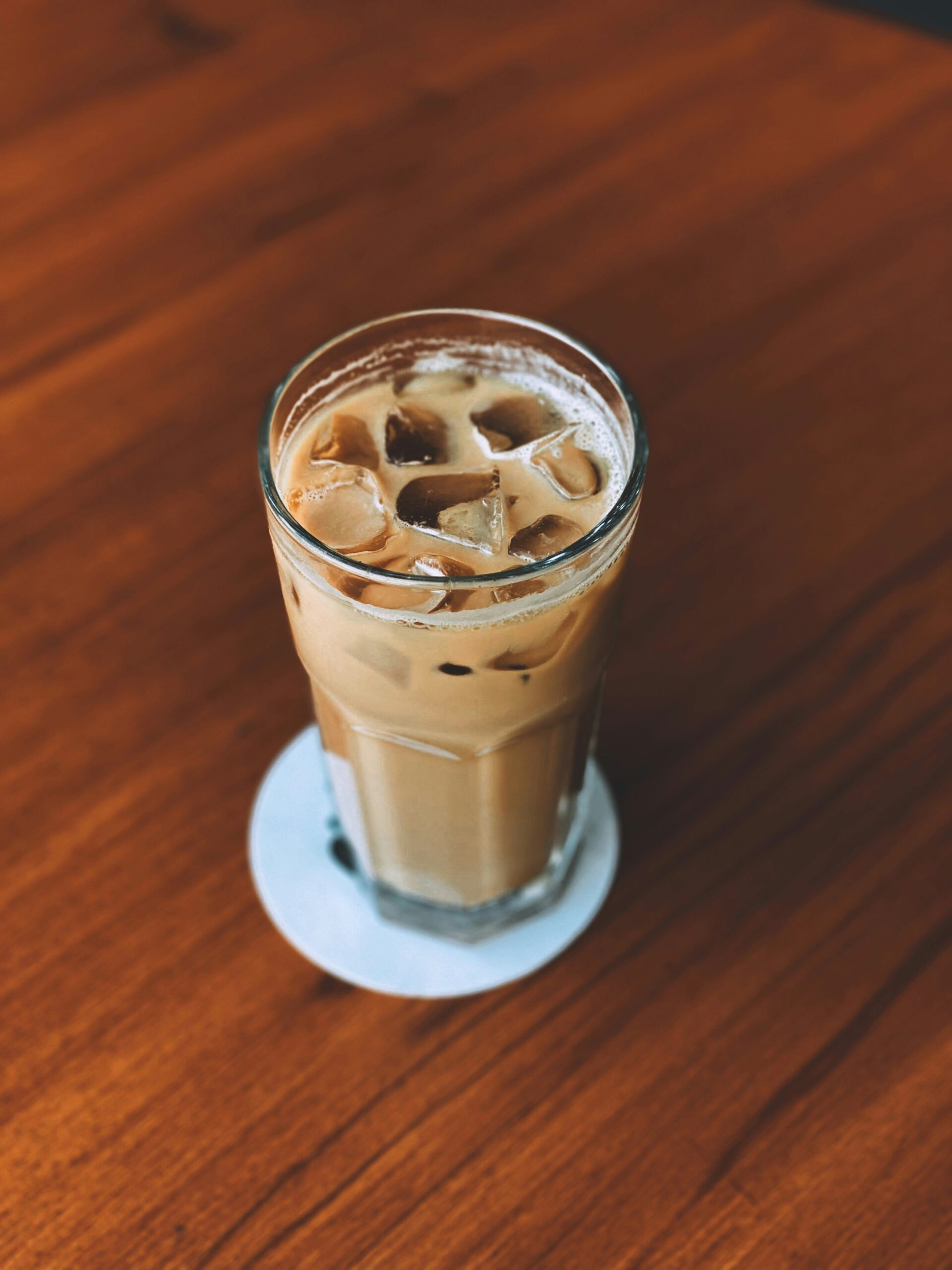 A glass of iced coffee with milk sits on a coaster on a wooden table.