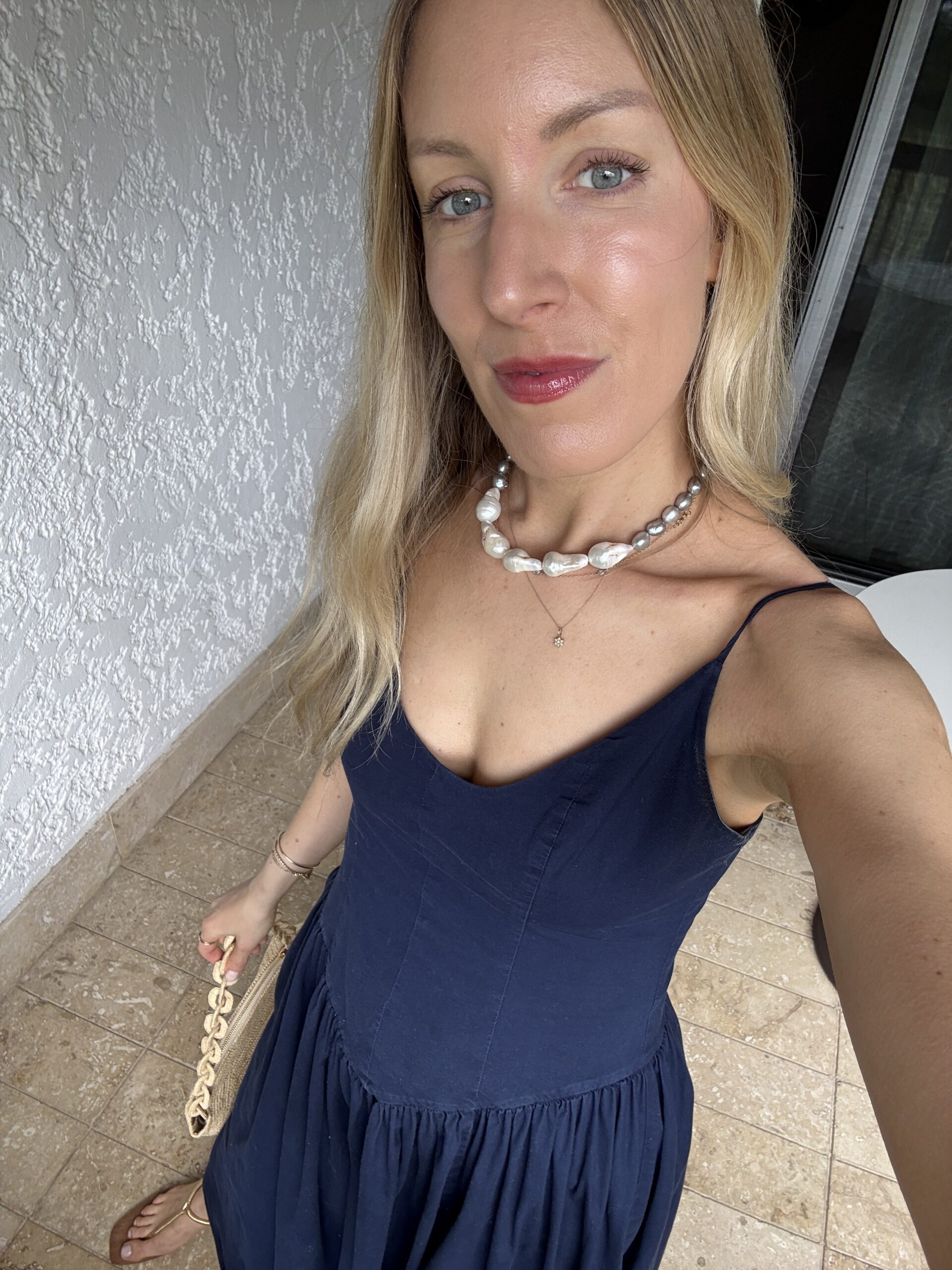 Person with long hair wearing a navy dress and a pearl necklace, standing on a tiled floor, holding a chain-strap handbag.